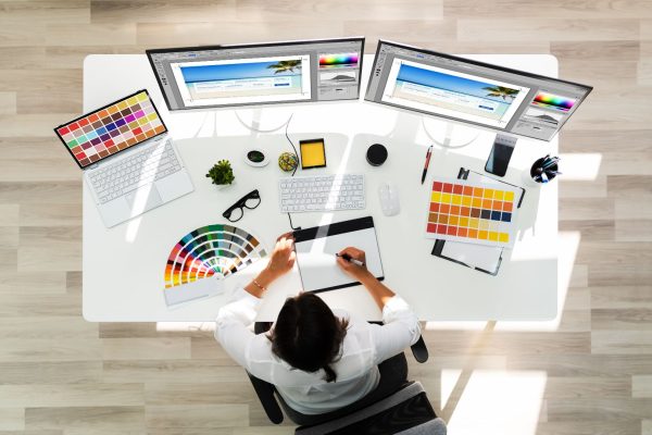 Mulher fazendo a construção de uma identidade visual. mesa com dois monitores e paletas de cores espalhadas.