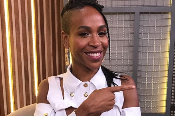 Karol Conka_e_uma_mulher_negra_veste_roupa_branca_usa_brincos_longos_e_esta_em_um_ambiente_fechado